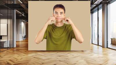 Close up studio portrait of young grimacing boy student wearing casual green t-shirt having fun pulling back his cheeks isolated on a beige background. People emotions, facial expression concept. Wall mural