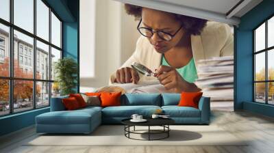 Closeup portrait young african american woman analyst expert with magnifying glass searching information in document sitting at desk with paper folder stack in comfort office. Research work Wall mural