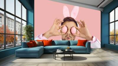 Closeup studio head shot of young man wearing white suit and cute plush toy bunny ears covering his eyes with two Easter eggs and making funny surprised face expression on pastel pink color background Wall mural