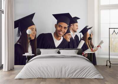 Confident in future. Smiling African-American exchange program student in black graduation cap and gown holding university diploma and looking at camera, standing together with international graduates Wall mural