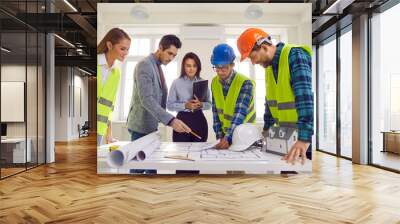 Construction team having discussion at work meeting. Group of multiracial male and female architects and builders wearing safety hard hats standing around office table looking at paper blueprint plans Wall mural