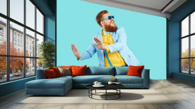 Cool cheerful stylish fat man rejoices and funny dances isolated on light blue background. Emotional red bearded chubby hipster man in good mood dressed in jacket, suspenders, jeans and sunglasses. Wall mural