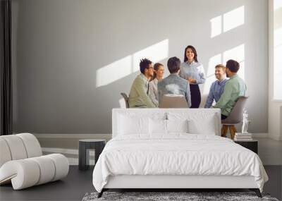 Corporate business team meeting with a manager or coach. Group of happy diverse, multiracial people sitting in a circle around a standing woman in an office with a light grey copy space wall Wall mural