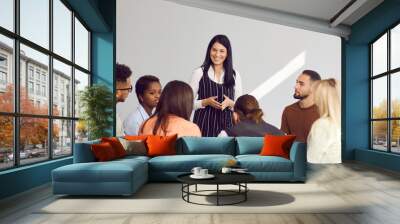 Corporate psychologist, team manager, motivational speaker or business coach meeting with diverse audience. Smiling beautiful woman standing among group of people and listening to questions they ask Wall mural