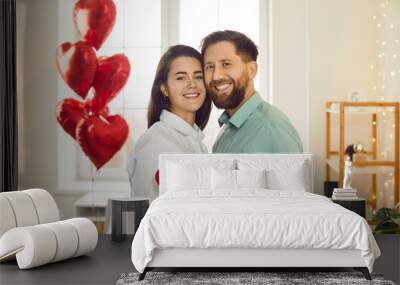 Couple in love celebrate St Valentine's Day and spend time together. Happy joyful man and woman standing in room decorated with lights and balloons, hugging, smiling and holding red heart shaped card Wall mural