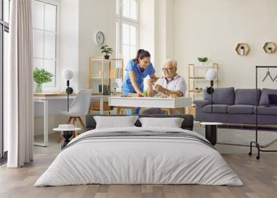 Demented senior man spends time in retirement home. Friendly nurse helping old man who is playing games and doing puzzles while sitting at desk in cozy retirement home interior. Dementia care concept Wall mural