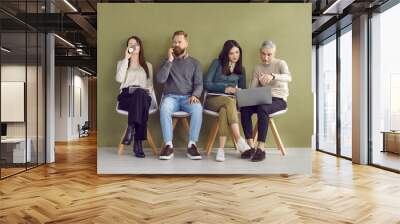 Different young and mature people sitting in line on row of chairs by green wall in company office, drinking coffee, using laptop, talking on phone, waiting for casual group meeting or job interview Wall mural