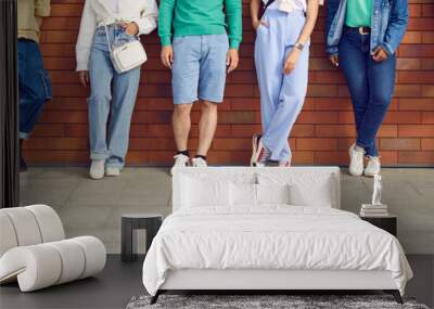 Diverse group of people in casual clothes. Six young multiethnic men and women in trendy jeans, shorts, and trainers leaning on brick wall in street. Cropped shot, human legs, street fashion banner Wall mural