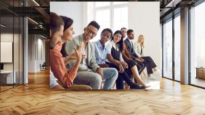 Diverse group of young people sitting in row in modern office and talking. Happy multi-racial men and women having interesting discussion during business meeting or psychological training Wall mural