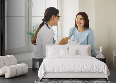 Doctor discussing treatment with cheerful smiley female patient. Happy physician and young woman talking and laughing sitting on examination bed in modern clinic or hospital Wall mural