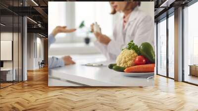 Doctor talking to patient and teaching her to take medicine, eat healthy food and stick to diet. Nutrition education and disease prevention concept. Blurred background, bowl of vegetables in close-up Wall mural