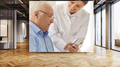 Doctor teaching senior patient to use healthcare app for setting medication reminder or tracking health indicators. Smiling nurse in assisted living facility showing photo of grandchildren to old man Wall mural