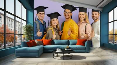 Education and success: Happy confident multiracial male and female university students, classmates and best friends in graduate hats give thumbs up standing together on purple color studio background Wall mural