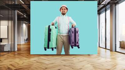 Embarrassed tourist guy holding two too heavy travel suitcases isolated on light blue background. Stylish man in panama and suspenders holds overweight luggage and looks at camera with funny grimace. Wall mural