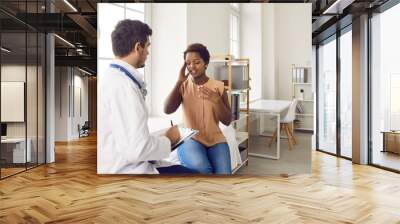 Embarrassed young dark-skinned woman tells doctor about headache during visit to clinic. Sick woman sits on examination couch and provides information about herself to doctor who writes on clipboard. Wall mural