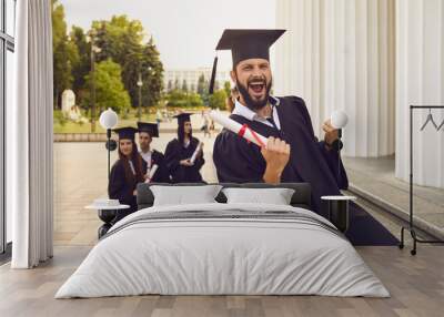 Emotional graduate wearing masters cap and gown raising up his hand with diploma. Brunette guy with a beard is ready for the next step in his life. Concept of success. Wall mural