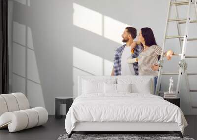 Family couple doing renovations at home. Young man and woman change color of walls in living room. Banner with happy husband and wife standing by ladder and looking at freshly painted light gray wall Wall mural