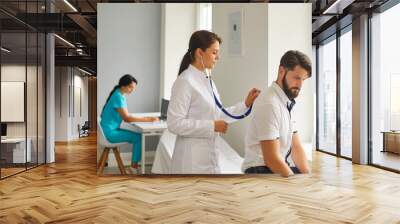 Female doctor listens to breathing with a stethoscope in a young patient in the hospital. Wall mural