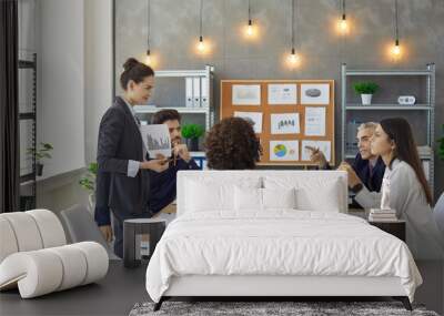 Female employee showing paper graph chat to colleague sitting at briefing desk. Businesspeople discussing project result, startup analysis report together in conference room during meeting at office Wall mural