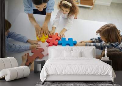 Female teacher engaging kids in teamwork. Little children participate in fun group activity in class. Small team of active students together joining big colorful jigsaw puzzle parts on classroom table Wall mural