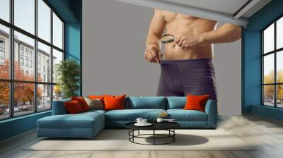 Fit man looks through magnifying glass in underpants at his private area. Shirtless attractive sexy guy in underwear briefs looking at crotch standing isolated on grey background. Penis size concept Wall mural