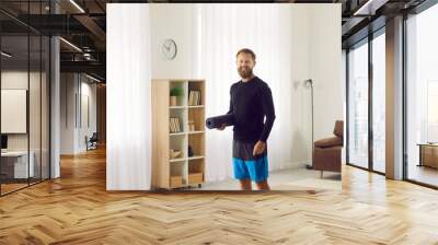 Fit young man having routine fitness and yoga workouts at home. Portrait of happy handsome guy with TWS earbuds standing in living room, holding exercise mat, looking at camera and smiling Wall mural