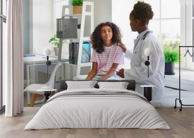 Friendly female pediatrician touches shoulder of teenage girl and informs her that there is no threat to her health. African American doctor and her preteen patient are talking in hospital office. Wall mural