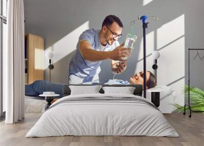 Friendly male nurse gives IV drip to happy female patient undergoing vitamin therapy. Smiling man in light blue scrubs sets intravenous line for cheerful young woman lying on bed in medical office  Wall mural