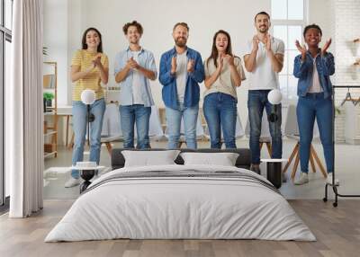 Friendly multiracial people in casual clothes stand in row and applaud looking at you. Group of friends in jeans and T-shirts joyfully clap their hands while standing in row in room. Team concept. Wall mural