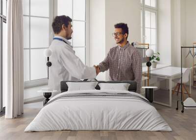 Friendly young male doctor shakes hands and greets patient in modern medical clinic. Young African-American man came for medical examination. Healthcare and professionalism concept. Wall mural