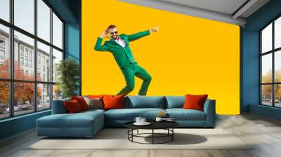 Full body happy funny smiling joyful handsome young man in trendy stylish green suit and sunglasses having fun, dancing isolated on yellow color background and pointing finger away at blank copy space Wall mural