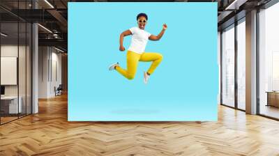 Full body length side profile view portrait happy excited Afro American woman in white tee, yellow pants and cool sunglasses jumping, running and flying high up in air on bright blue studio background Wall mural