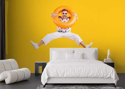 Full body photo of a happy funny man in sunglasses holding rubber ring and jumping isolated on studio yellow background. Funny tourist is going on summer holiday trip. Vacation and travel concept. Wall mural