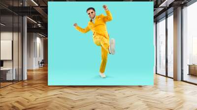 Full body shot of a happy, cheerful man wearing a trendy, stylish yellow suit and sunglasses having fun and dancing on a turquoise blue studio background. Party and fashion concept Wall mural