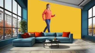 Full body studio shot of a cheerful smiling girl wearing orange sweatshirt and blue jeans. Happy young woman in a comfortable casual outfit walking forward against a vibrant yellow colour background Wall mural