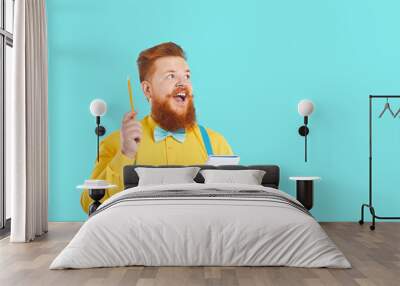 Funny cheerful bearded man in yellow shirt and blue bow tie standing on blank copyspace background, holding pencil and paper, thinking of cool brand name, writing brilliant ideas for marketing slogan Wall mural