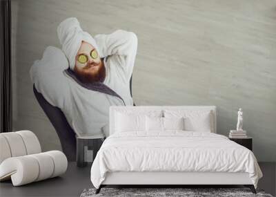 Funny fat bearded man with a cosmetic mask on his face in bathrobe towel on his head on his face resting against a gray background Wall mural