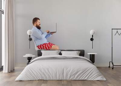 Funny fat man in a blue shirt and red shorts sitting on a chair working online using a laptop on a gray background. Wall mural