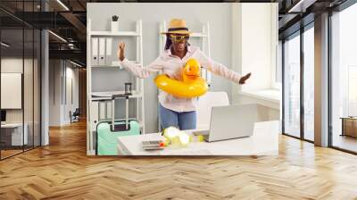 Funny happy african american woman standing at the desk on workplace at office having fun in rubber ring with suitcase and booking tickets for summer vacation online via laptop. Holiday trip concept. Wall mural