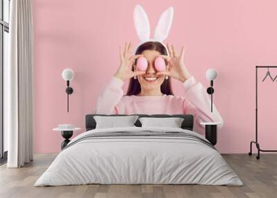 Funny joyful brunette young woman in pink sweatshirt with bunny ears on hair band holding easter eggs on eyes on pink background. Happy girl celebrating easter in rabbit costume with pink earrings. Wall mural