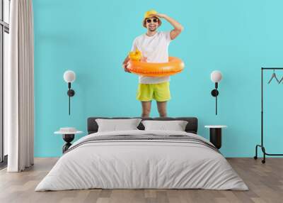 Funny joyful young man set up for summer vacation and looking for good offer from travel agency. Happy guy in summer clothes and with inflatable circle looking into distance on light blue background. Wall mural