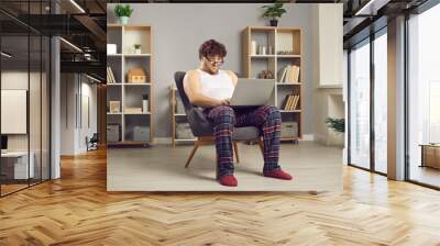 Funny serious fat man in pajamas and eyewear looking at screen working online on laptop from home sitting on chair in apartment living-room. Charismatic curly overweight guy uses internet Wall mural