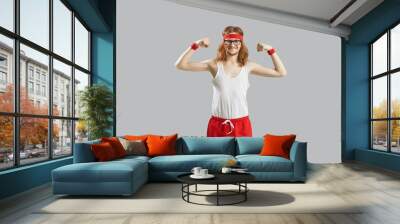 Funny thin skinny man with long hair in eyeglasses, retro headband, white tank top and red shorts showing weak muscles standing isolated on gray background. Sports workout and fitness exercise concept Wall mural