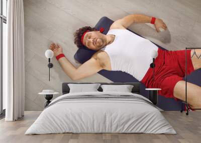 Funny tired fat man lying on fitness mat after gym workout. Exhausted guy in retro sweatband, tank top and shorts fallen on floor after sports exercise with dumbbells. Top view, high angle, from above Wall mural