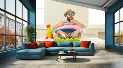 Funny young man in sunglasses and summer wear sipping beach cocktail, sitting barefoot in inflatable lifebuoy at home. Covid-19 quarantine, vacation in lockdown, canceled holiday travel plans concept Wall mural