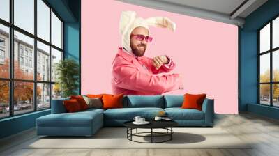 Goofy guy in funny rabbit costume having fun in the studio. Happy confident bearded young man wearing suit, plush Easter bunny ears and pink party glasses dancing isolated on pink colour background Wall mural