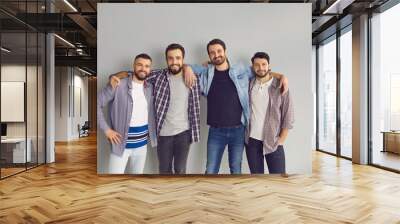 Group of cheerful friends having fun together. Portrait of happy bearded young men standing with arms around each other's shoulders and smiling at camera in studio with light gray background Wall mural