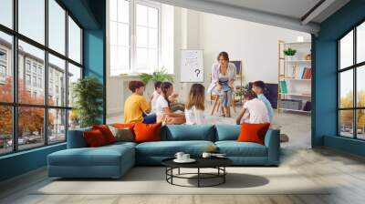 Group of elementary school students sitting on floor listening to teacher in classroom. School children sitting cross legged in circle around smiling female teacher listening attentively Wall mural