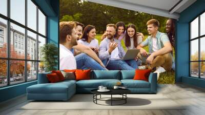 Group of happy, cheerful, diverse, multiethnic student friends sitting on green grass outside, studying information for project together, discussing something, using online tools on modern laptop PC Wall mural