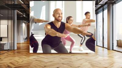 Group of happy, positive, smiling young people having a fitness workout at the gym. Cheerful sporty well trained men and women in comfortable sportswear doing a squats exercise together Wall mural